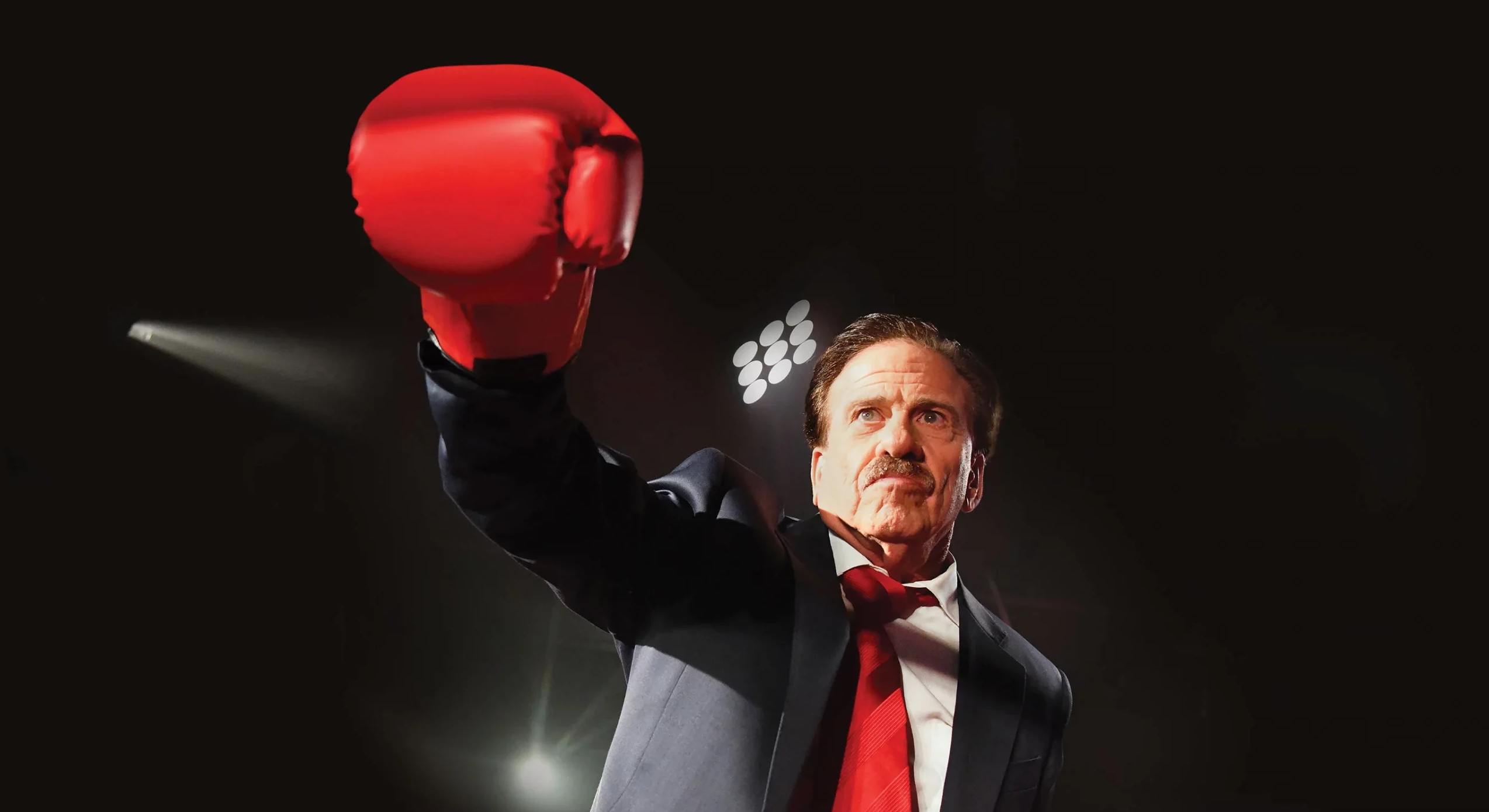 A man in a suit holds up his right hand which has a boxing glove