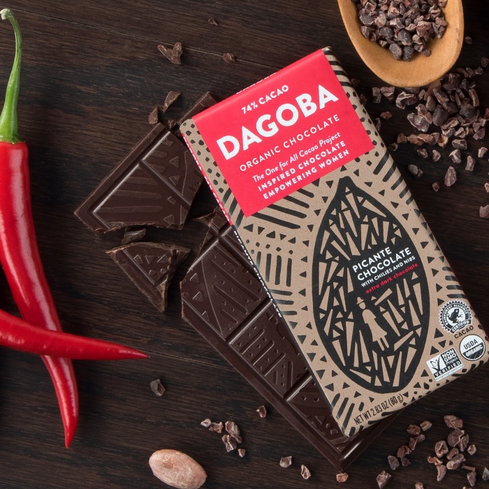 Dagoba chocolate bar on a wood table next to cacao nibs and chili peppers