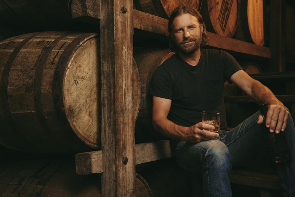 Dierks Bentley seated beside bourbon barrels with a glass of ROW 94 whiskey