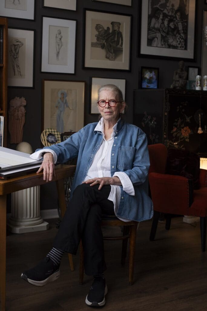 Illustrator Claudia Hammer seated in her home studio in a denim shirt, surrounded by framed artwork
