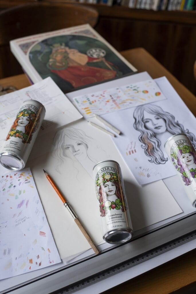 Greater Than cans resting on a drafting table beside pencil sketches and color swatches of the goddess illustration